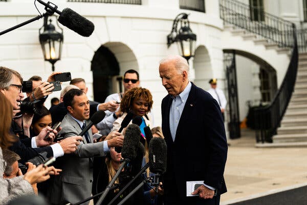 President Biden, wearing a dark jacket and blue shirt, stands in front of people holding microphones and cameras.