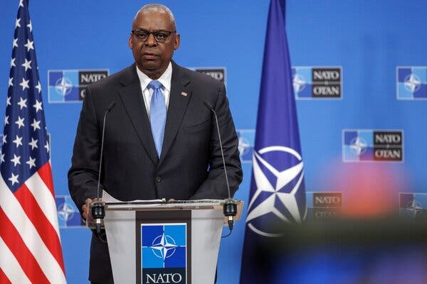 Lloyd Austin stands at a podium wearing a suit and tie. There are flags to his right and left.