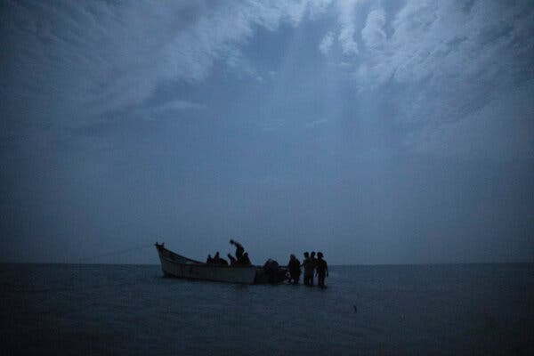 A blurry, dark picture of a boat in water. People in silhouette are boarding or standing nearby. 