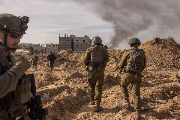 Soldiers walk across a stretch of dirt and debris, with buildings and a plume of smoke in the background.