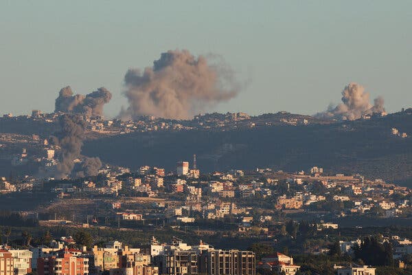 Gray smoke billows up from behind a green hill, with a city in the foreground. 