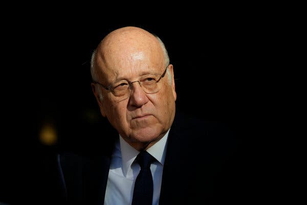 A portrait of Najib Mikati wearing glasses, a blue shirt and dark tie and suit.