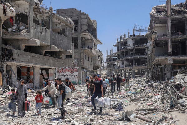 People walk through a road littered with debris. Bombed out buildings rise on either side of them. 