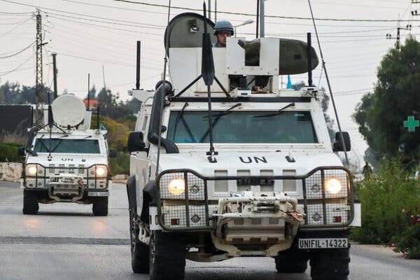 Two white military-style vehicles drive down a street.