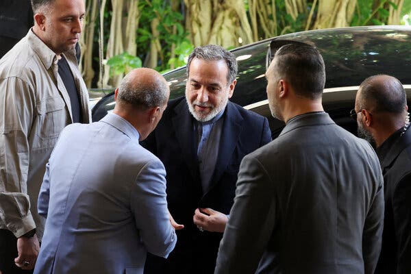 A bearded man in a dark suit is talking to another man with his back to the camera outside a black car.