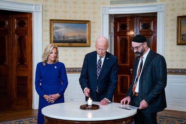 President Joe Biden lights a candle on a table while standing next to Jill Biden and Rabbi Aaron Alexander.