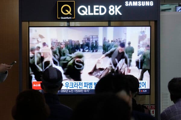 People sit in front of a large-screen television showing soldiers in green fatigues standing in line.