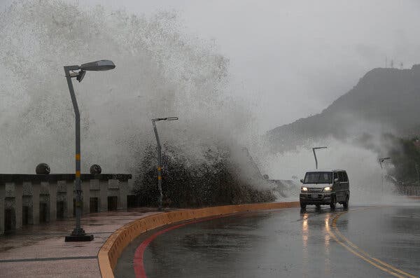 A vehicle moves along a road as a wave crashes over a barrier.