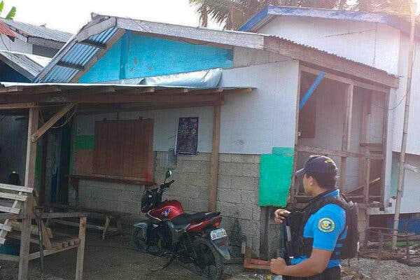 A police officer stands in front of a small house made of concrete blocks, with a motorcycle in front of it. 