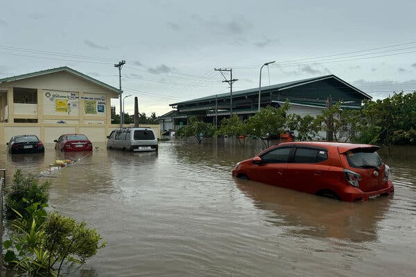 Four cars are partially submerged in brown floodwaters outside a yellow building. 