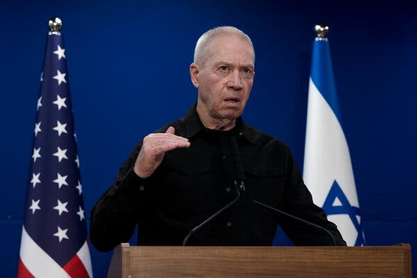 Yoav Gallant gestures while standing at a lectern, flanked by an Israeli flag and American flag. 