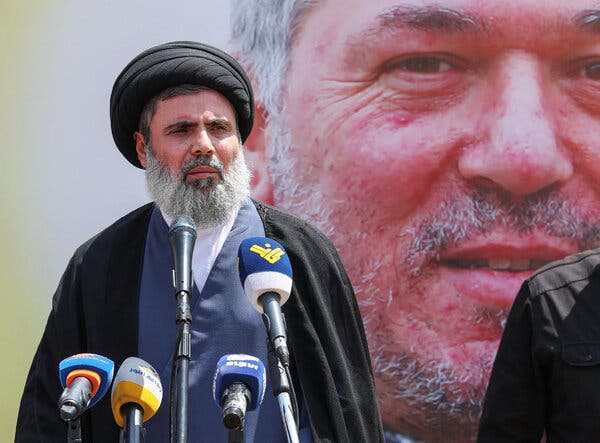 Hashem Safieddine, wearing a black turban and a black robe, stands in front of microphones. A large image of a man’s face is shown behind him.