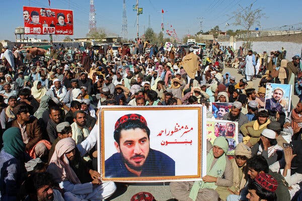 A poster bearing the image of poster of Manzoor Pashteen is held up by men sitting amid a crowd.