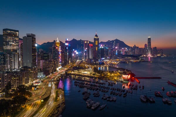 An urban, waterfront landscape at sunset shows brightly lit, massive skyscrapers looking over a marina and a waterside highway bustling with vehicles.