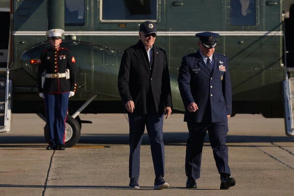 President Biden, wearing a baseball cap, is on a tarmac, walking with a decorated military man; behind him is a dark green aircraft and a soldier standing at attention.