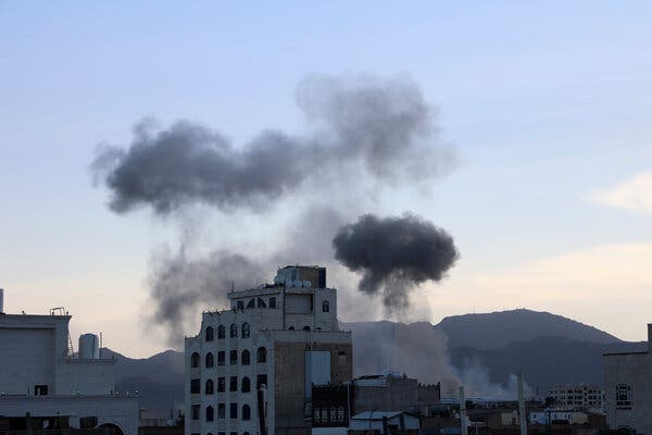 Smoke rises behind a damaged building,  with mountains in the background. 