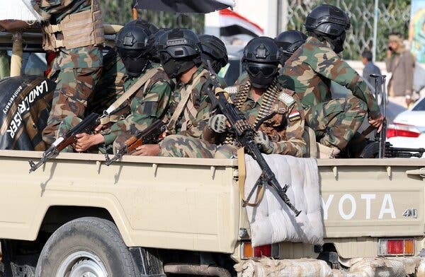 People in fatigues wearing helmets and carrying guns in the back of a pickup truck.