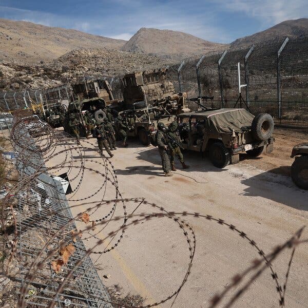 Military vehicles and soldiers move on a dirt road in between barbed-wire fences, with barren hills in the background.