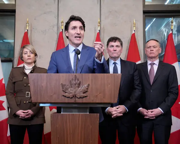 Prime Minister Justin Trudeau raises his hand as he speaks at the podium. Three people stand behind him in the background, and behind them are several Canadian flags.