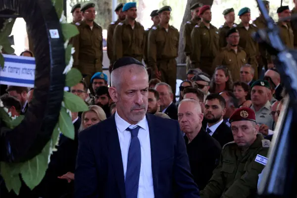 A man in a suit stands in front of a seated crowd and a number of security personnel.