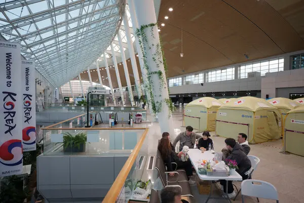 People sit at a table in a high-ceilinged airport terminal building with yellow tents set up behind them.