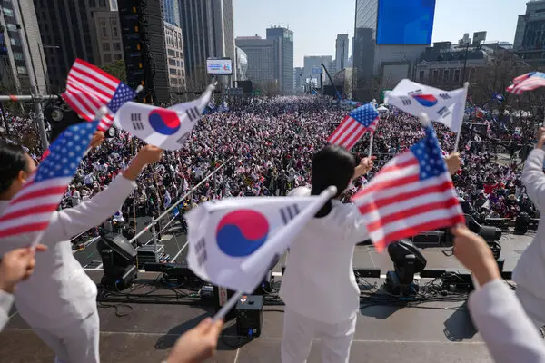 A huge crowd stands in a large open area. In the foreground, people wave South Korean and American flags.