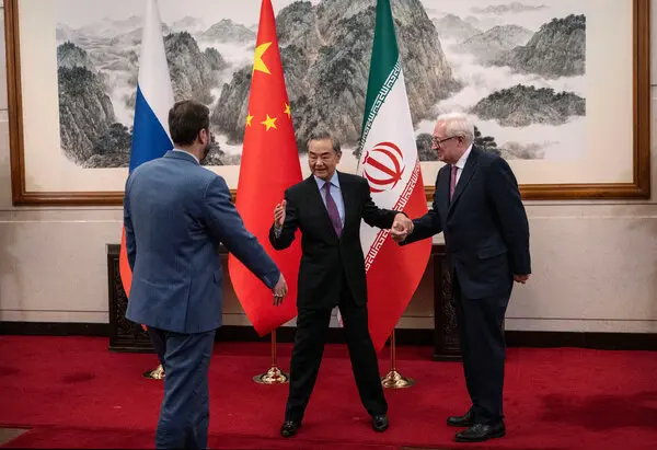 Three men in suits stand in a room with a red carpet, behind them are the flags of Russia, China and Iran.