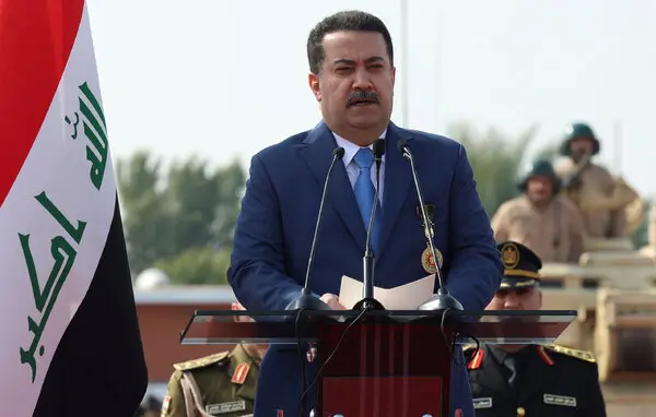 Iraqi Prime Minister Mohammed Shia al-Sudani speaks into microphones at a lectern next to the Iraqi flag.