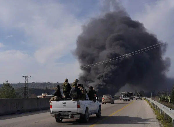 Smoke rises as Syrian troops drive down the road in the back of a pickup truck.