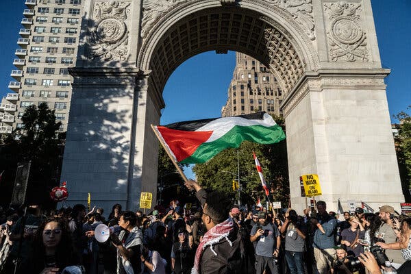 Protesters carrying signs and a Palestinian flag gather under the Washington Arch on a sunny day.