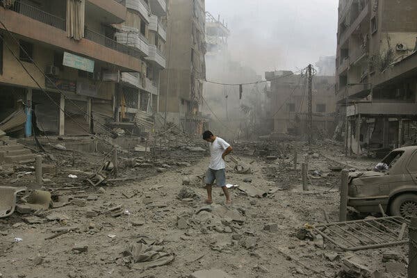 A person in a white T-shirt and blue shorts stands amid rubble and damaged buildings. Objects, including a car, appear covered in dust.