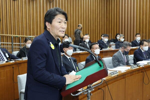 A man standing at a lectern and speaking. Behind him are two curved rows of desks.