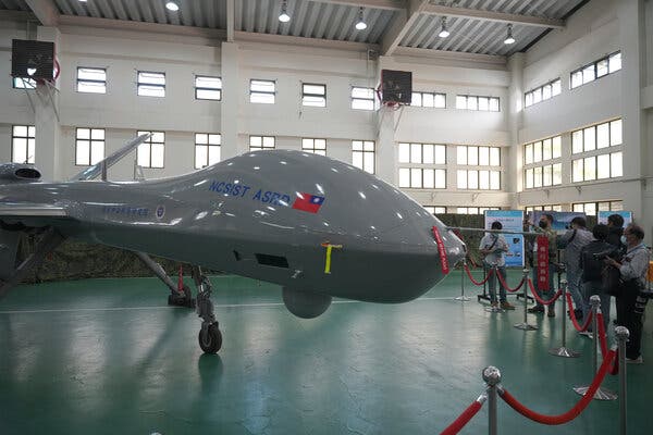 A large gray military drone, with the flag of Taiwan painted on it, is on display inside a large building. It is cordoned off by a red rope, and people are standing behind it, taking pictures. 