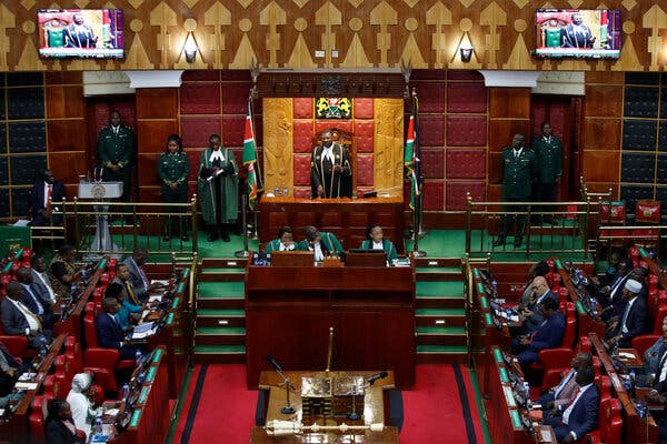 People sit or stand inside a legislature as a person addresses the room from a central podium.