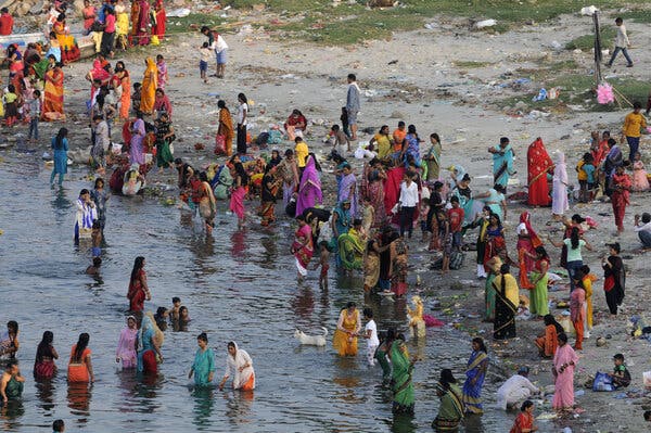 A riverbank is crowded with people, many of them women in colorful saris. Some people are in the water.