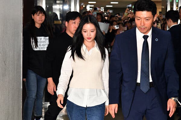 A woman in a white shirt, white sweater and bluejeans walking next to a man in a blue suit. A throng of photographers is behind them.