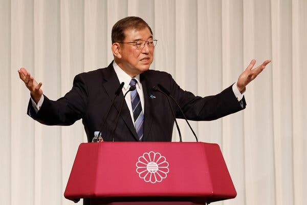 Shigeru Ishiba, standing behind a red lectern with four thin microphones, addressing a news conference, his arms outstretched. 