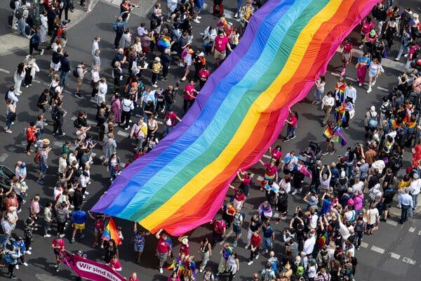 A large crowd carries a long rainbow striped flag down the middle of a street during a Pride parade.