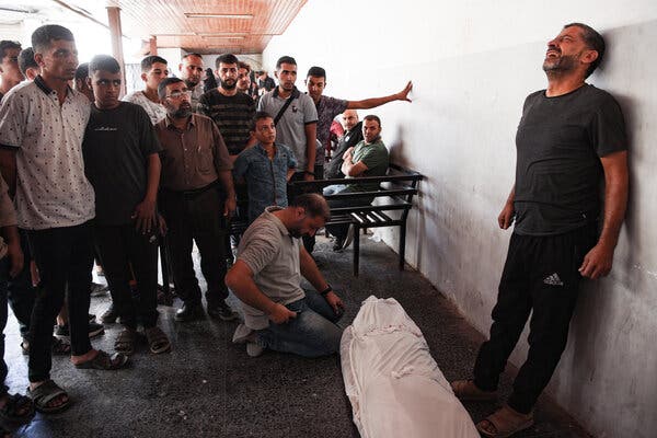 In a hospital, a man leans, crying, against a wall, next to a shrouded body, while others look on.