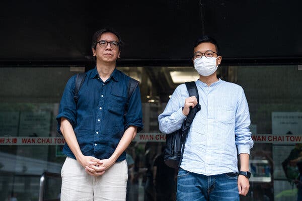 Two casually dressed men with backpacks  stand in front of a building.