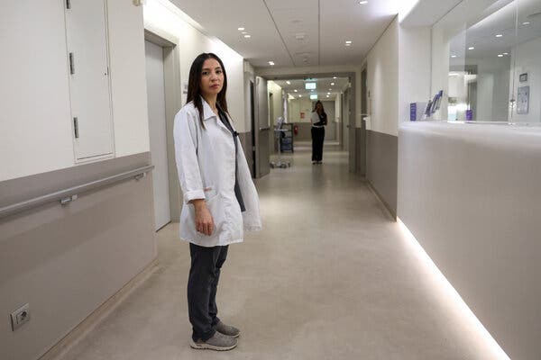 A woman stands in an empty hallway wearing a white coat. She is looking at the camera. 