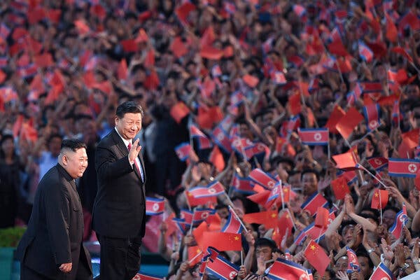 President Xi Jinping of China, standing beside the North Korean leader Kim Jong Un, waves to a crowd of people holding Chinese and North Korean flags.