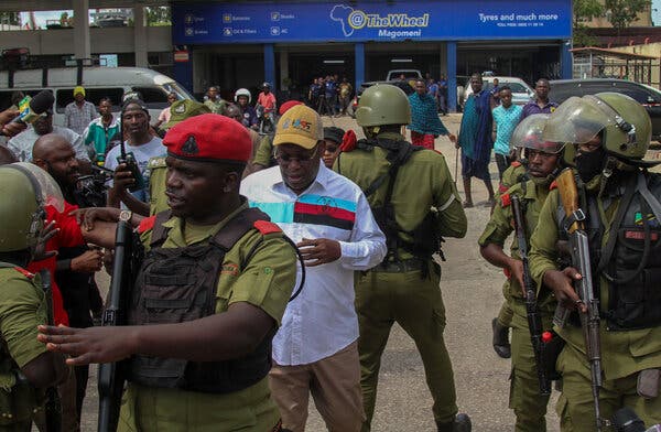 Police officers wearing helmets and protective gear surround a man wearing a white shirt and a yellow cap.