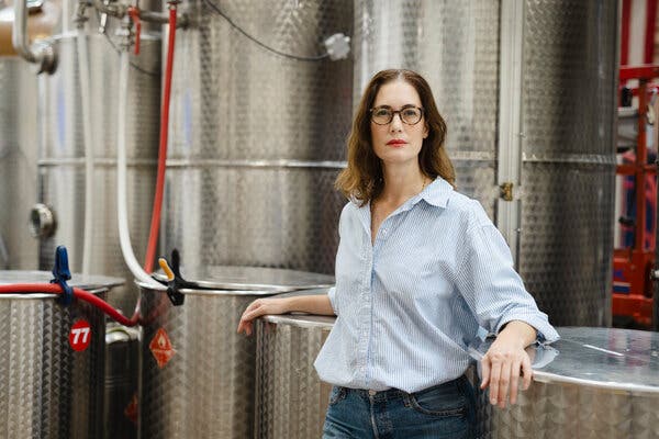 Sonat Birnecker Hart, wearing a collared shirt and jeans, stands in front of a line of large silver tanks with red pipes running through them.